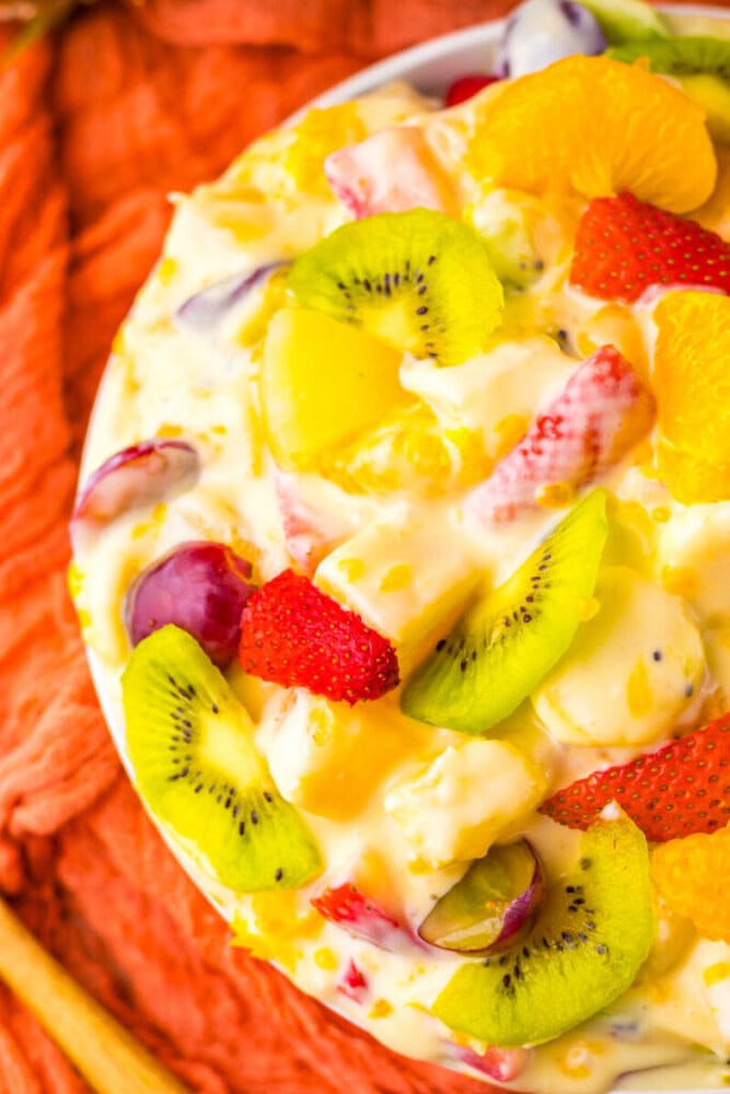 A close-up of a fruit salad with kiwi, strawberries, grapes, orange slices, and pineapple pieces mixed in a creamy dressing, served in a white bowl on an orange cloth.