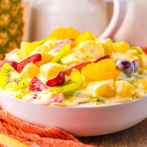 A white bowl filled with creamy fruit salad containing pineapple, grapes, orange segments, strawberries, and kiwi slices, set on a wooden surface with an orange cloth.