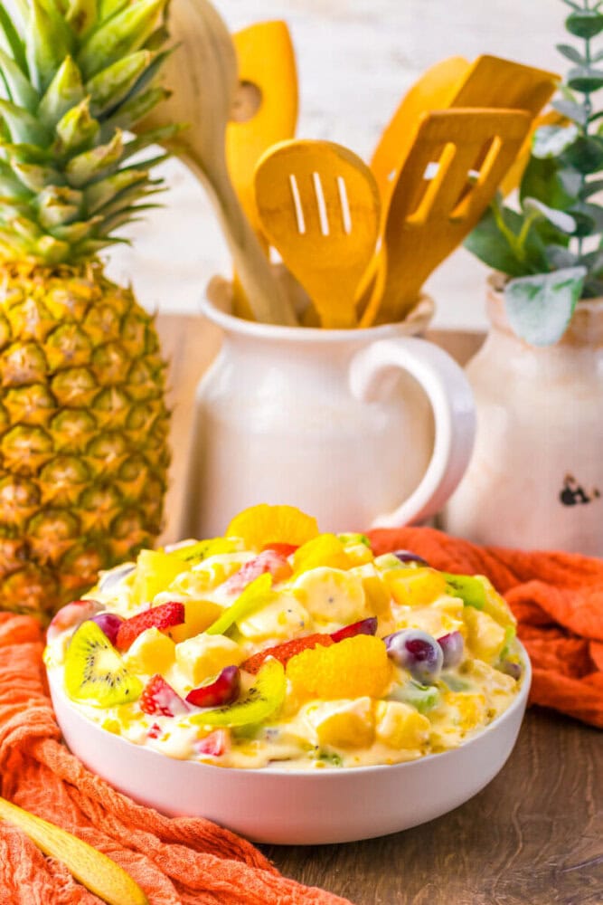 A bowl of creamy fruit salad with pineapple, kiwi, strawberries, grapes, and orange slices sits on a table next to a pineapple, wooden utensils, and ceramic jars.
