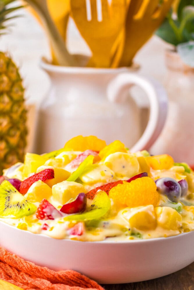 A bowl of creamy fruit salad containing pieces of kiwi, strawberries, grapes, pineapple, and orange segments sits on a table with a pitcher and utensils in the background.