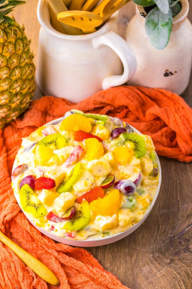 A bowl of creamy fruit salad with sliced kiwi, grapes, mandarin oranges, strawberries, and pineapple on a wooden table with a pineapple, jars, and an orange cloth nearby.