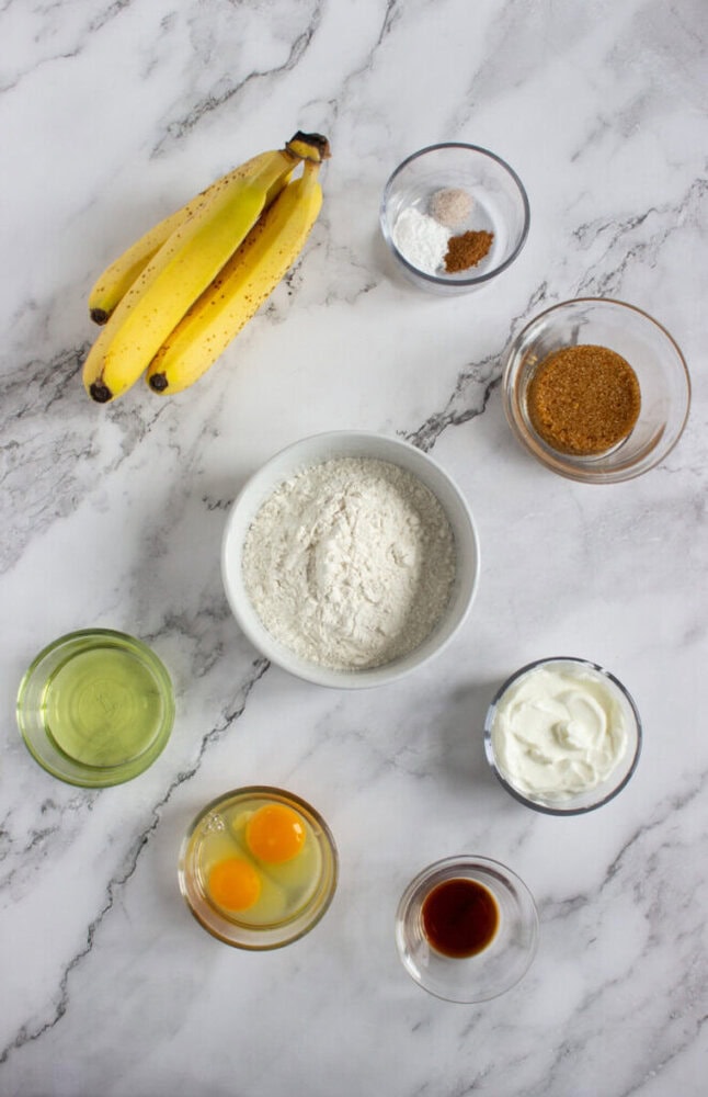 Top-down view of ingredients for baking: bananas, flour, eggs, oil, yogurt, brown sugar, vanilla extract, baking powder, baking soda, cinnamon, and nutmeg on a marble surface.