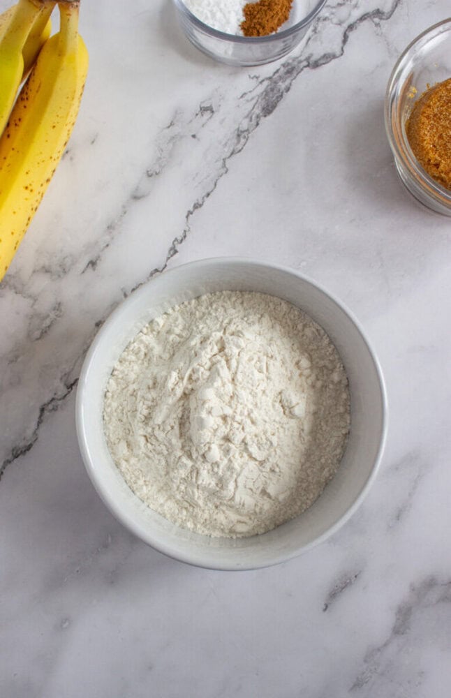 A white bowl filled with flour sits on a marble countertop beside bananas and small bowls of spices.