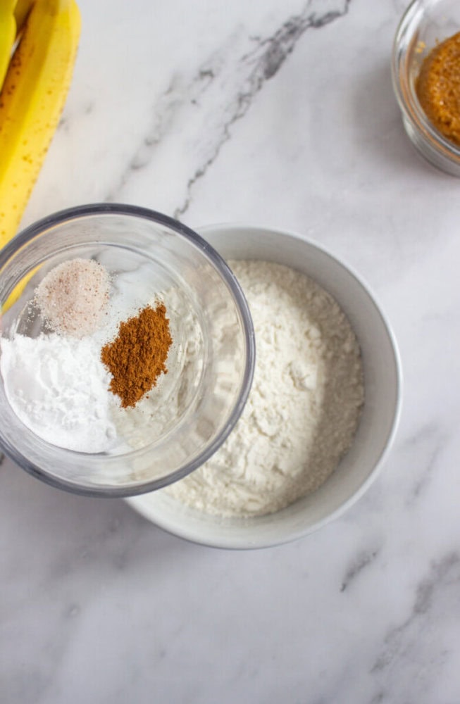 A glass bowl with baking soda, salt, and cinnamon above a white bowl of flour on a marble countertop, with part of a banana and another bowl in the background.