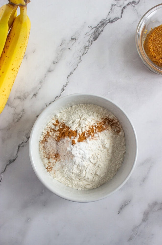 A white bowl with flour, baking soda, salt, and cinnamon sits on a marble countertop next to bananas and a small bowl of brown sugar.