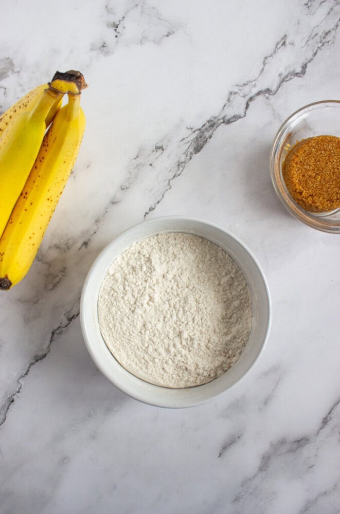 A bowl of flour, a bunch of ripe bananas, and a small bowl of brown sugar sit on a white marble surface.