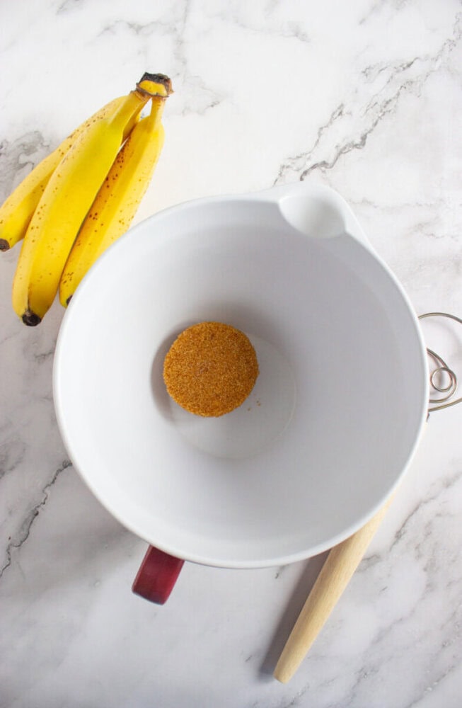 A bunch of bananas sits next to a white mixing bowl containing brown sugar on a marble countertop, with a wooden spoon nearby.