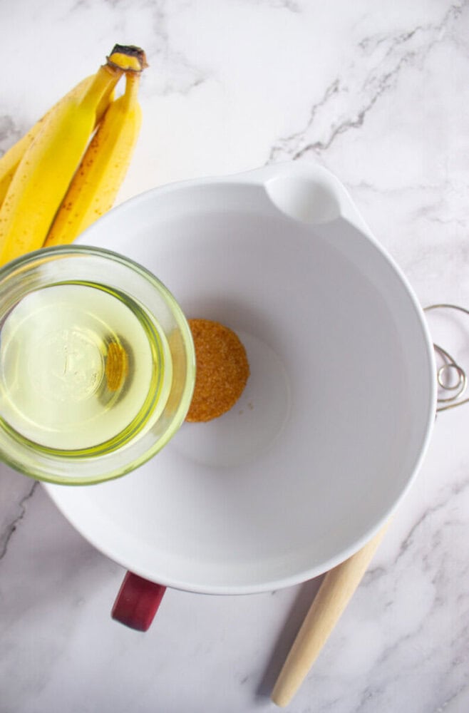 A bunch of bananas, a white mixing bowl with sugar, a glass of oil being poured in, and a wooden spoon on a marble surface.