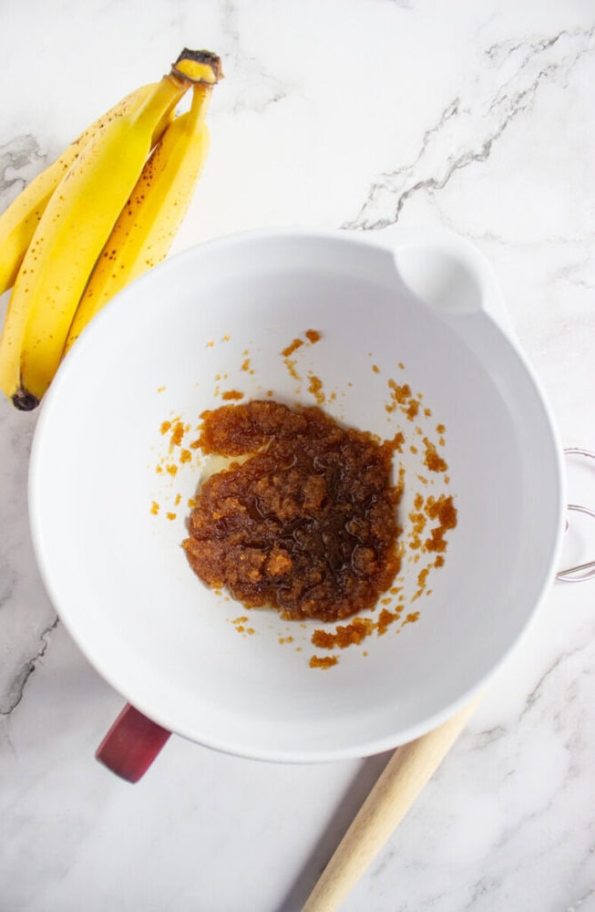 A white mixing bowl with mashed brown sugar and butter sits on a marble surface next to ripe bananas and a wooden-handled whisk.