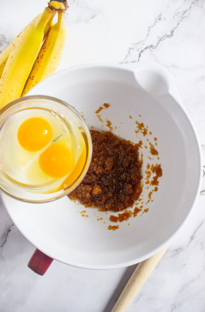 A mixing bowl with brown sugar and butter mixture, a glass bowl with two eggs above it, and two ripe bananas on a marble countertop.