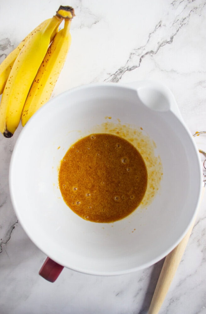 A white mixing bowl with a yellow-brown liquid mixture inside is on a marble surface next to a bunch of ripe bananas.