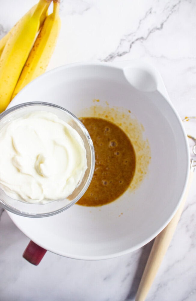 A bowl with a brown mixture, a glass containing yogurt, bananas nearby, and a whisk on a marble surface.