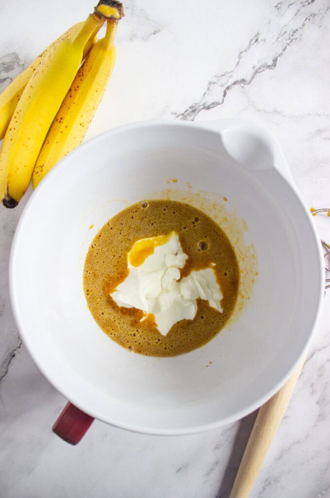 A mixing bowl with a batter and a dollop of yogurt, next to a bunch of ripe bananas on a marble countertop.