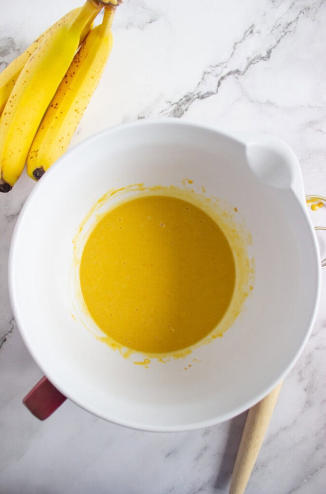 A white mixing bowl with yellow batter inside sits on a marble surface next to a bunch of bananas and a wooden-handled whisk.