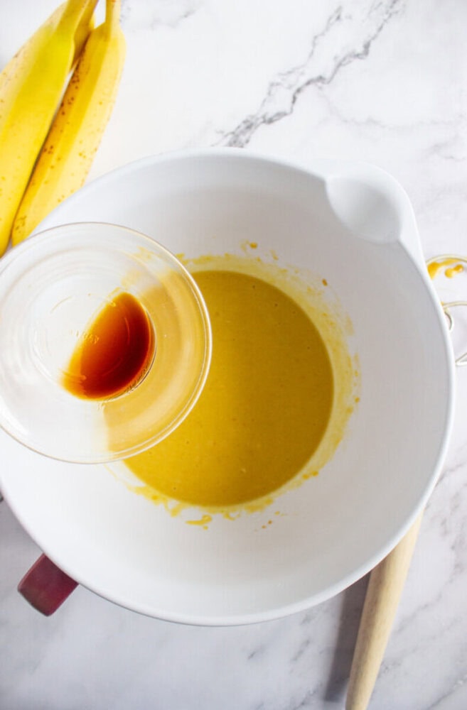 A glass bowl of vanilla extract is being held over a mixing bowl containing a yellow batter, with bananas and a wooden spoon nearby on a marble surface.