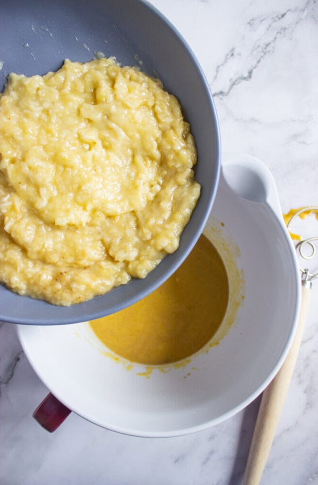 Mashed bananas are being poured from a gray bowl into a white bowl containing a yellow batter mixture on a marble surface.