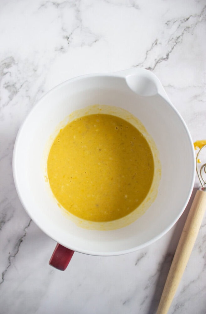 A white mixing bowl containing a yellow batter sits on a marble countertop next to a whisk with some batter on it.