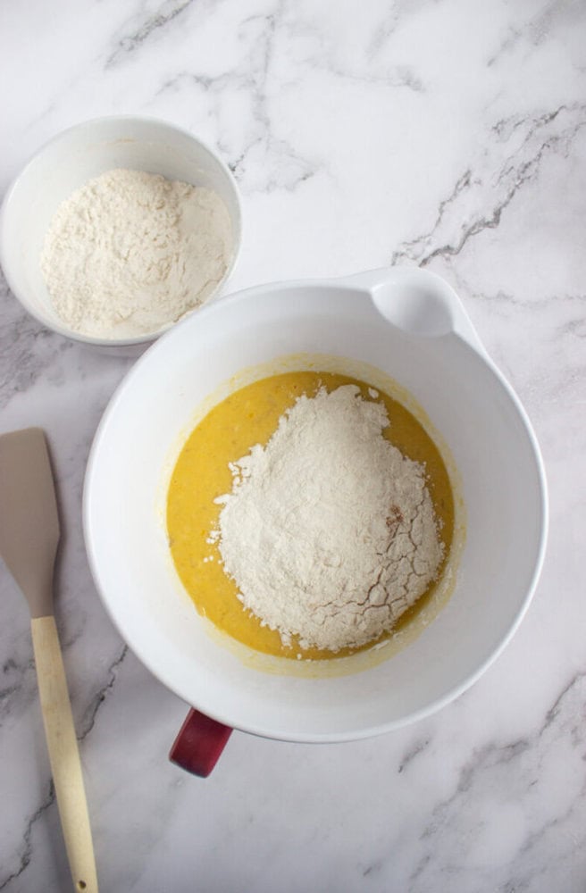 A mixing bowl with flour and a yellow batter, a spatula, and a smaller bowl of flour on a marble countertop.