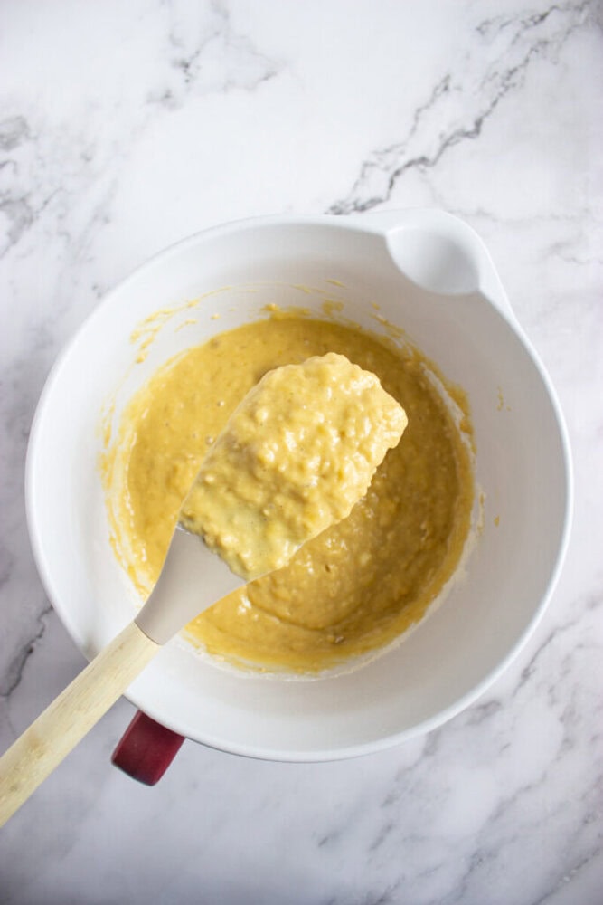 A white mixing bowl containing yellow batter with a spatula resting on the edge, set on a marble countertop.