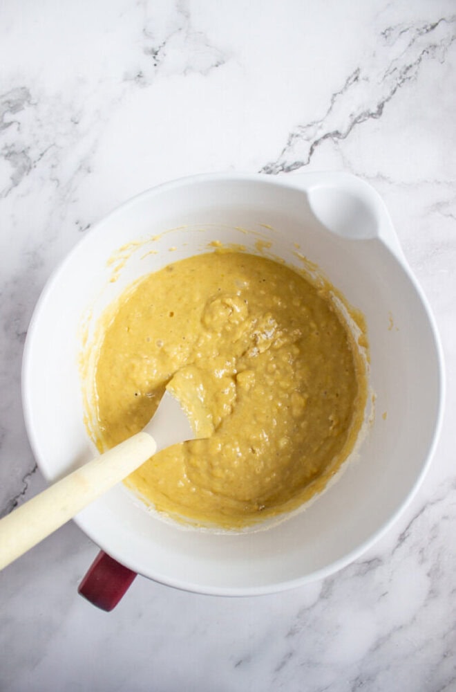A white mixing bowl with yellow batter and a spatula resting inside, placed on a white marble surface.