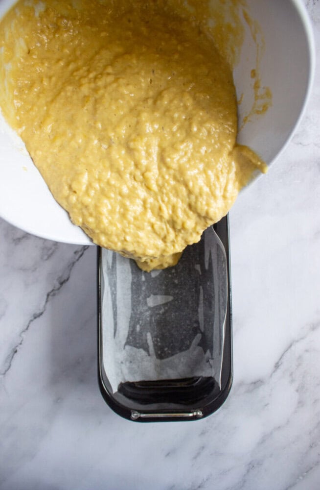 A bowl of banana bread batter is being poured into a parchment-lined loaf pan on a marble countertop.