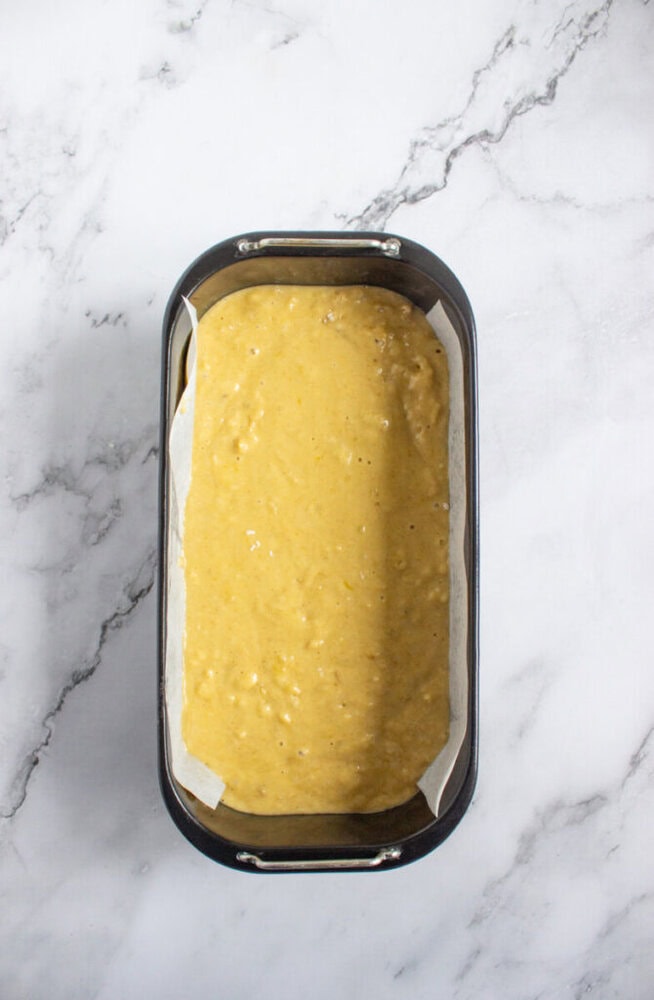 A loaf pan lined with parchment paper is filled with uncooked banana bread batter, resting on a white marble surface.