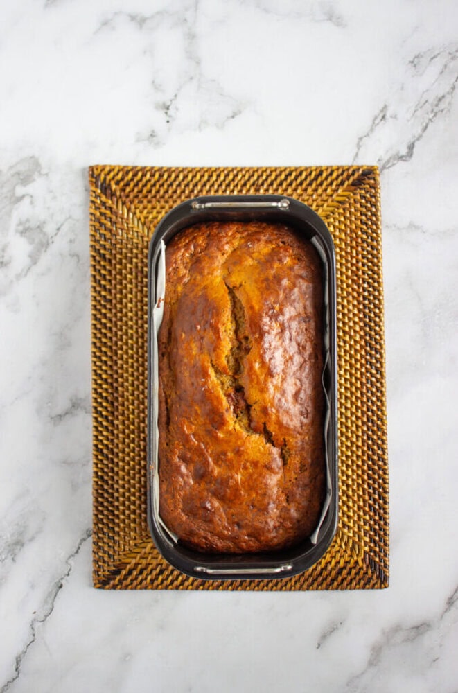 A loaf of banana bread in a metal baking pan sits on a woven placemat atop a white marble surface.