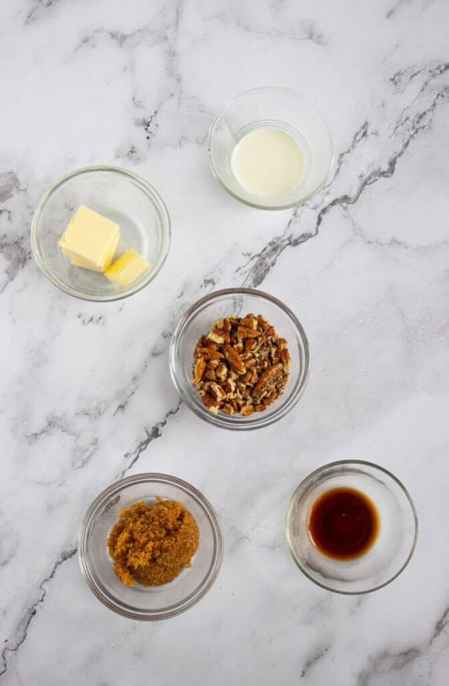 Five small glass bowls containing butter, milk, chopped pecans, brown sugar, and vanilla extract arranged on a marble countertop.