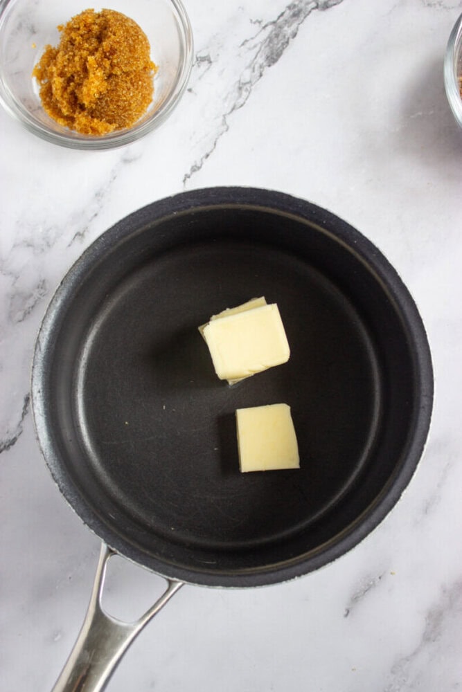 A saucepan with two cubes of butter inside, placed on a marble countertop next to a small bowl of brown sugar.