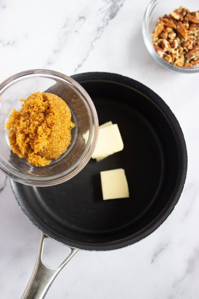 A saucepan with two pieces of butter inside, a small bowl of brown sugar held above it, and a separate bowl of chopped pecans on a marble countertop.