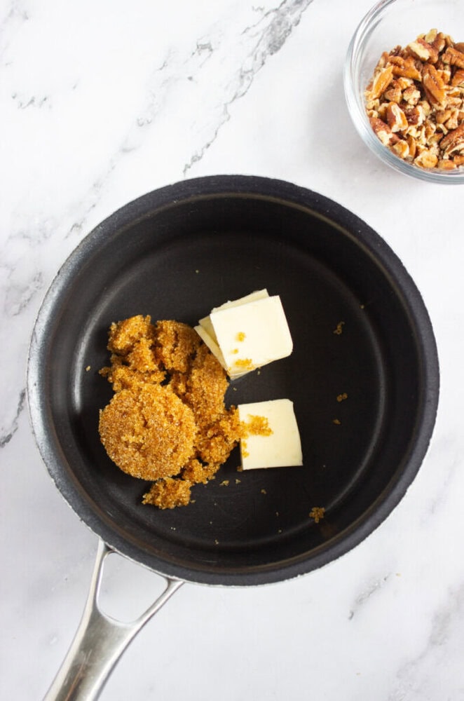 A saucepan containing brown sugar and butter sits on a white surface next to a small glass bowl of chopped pecans.