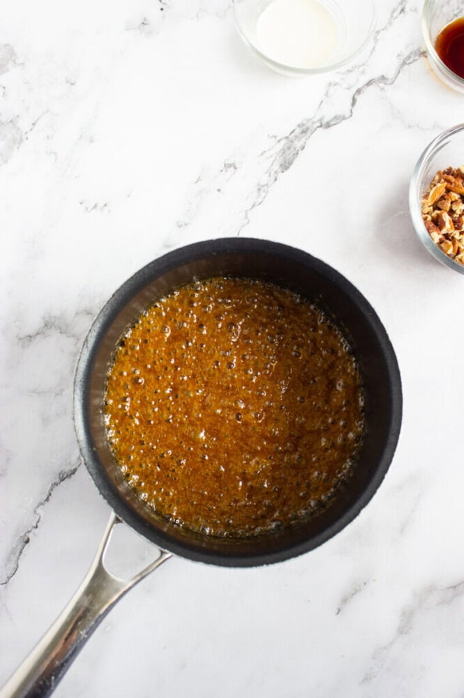 A saucepan with bubbling caramel mixture sits on a marble countertop, surrounded by small bowls containing cream, vanilla extract, and chopped pecans.