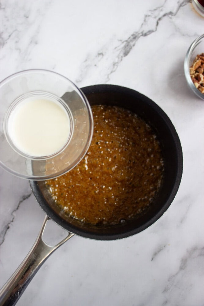 A saucepan with bubbling brown mixture sits on a marble surface, while a glass bowl of milk is held above it, ready to pour in. A small bowl of pecans is nearby.