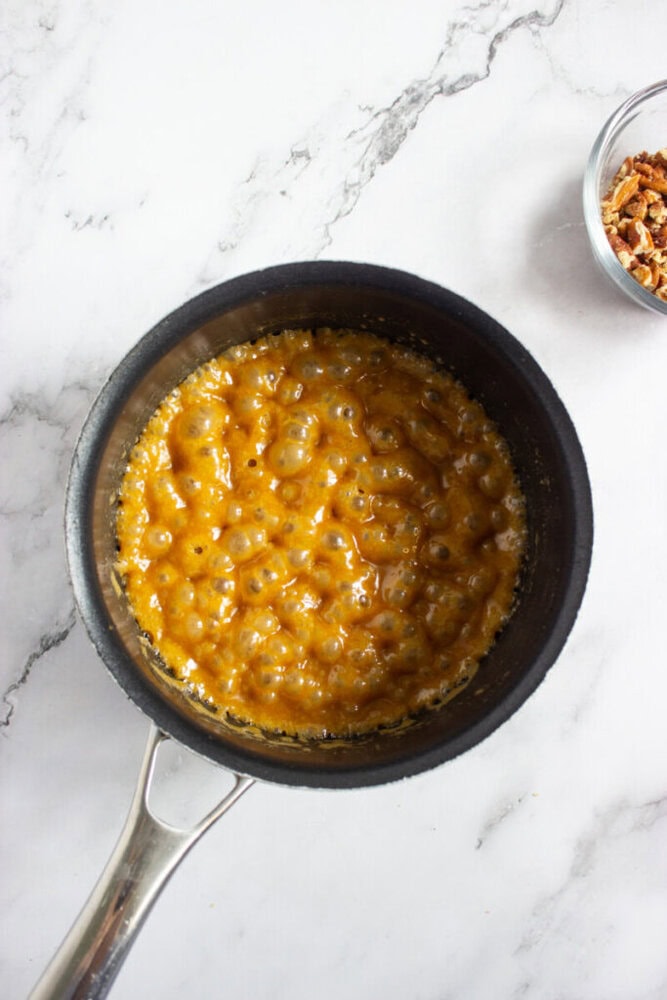 A saucepan filled with bubbling caramel mixture sits on a marble surface, with a small bowl of chopped pecans nearby.