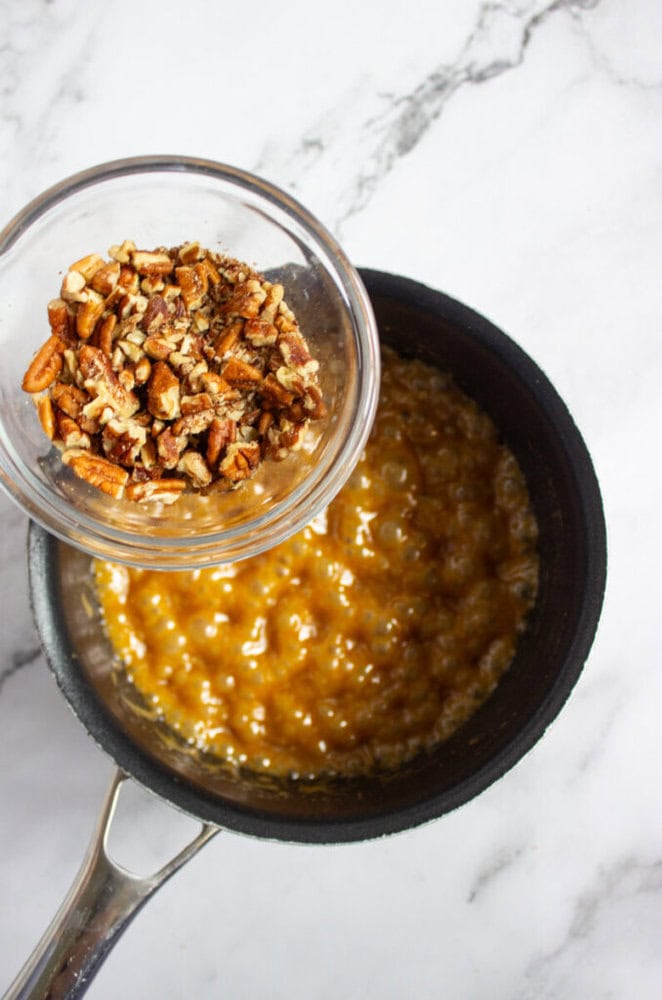 A glass bowl of chopped pecans is being poured into a saucepan of bubbling caramel mixture on a marble countertop.