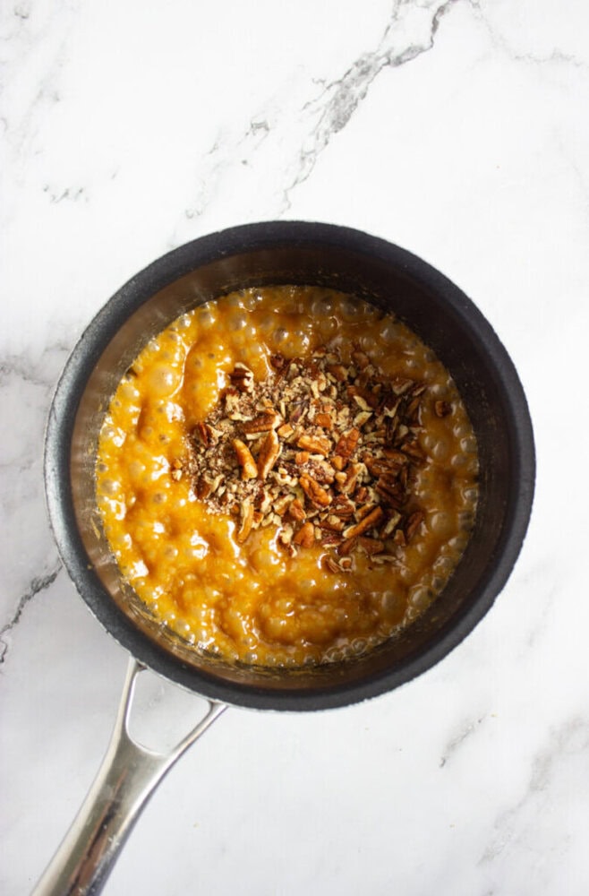 A saucepan filled with bubbling caramel mixture topped with chopped pecans sits on a white marble surface.