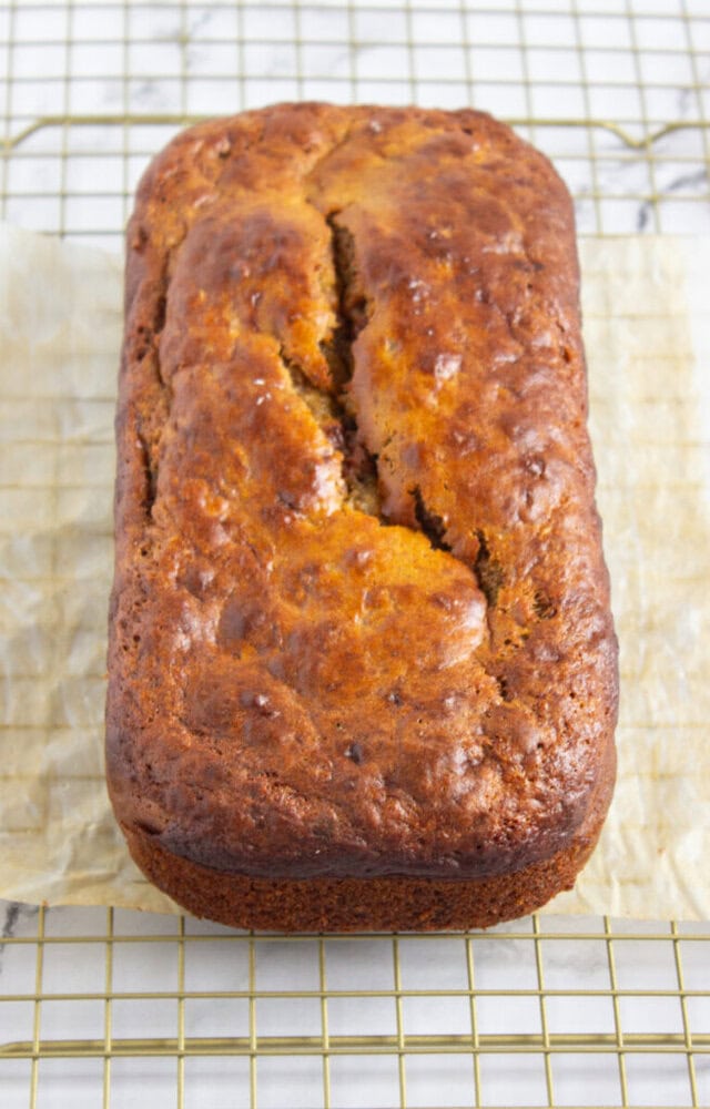 A loaf of golden-brown banana bread sits on parchment paper atop a wire cooling rack.