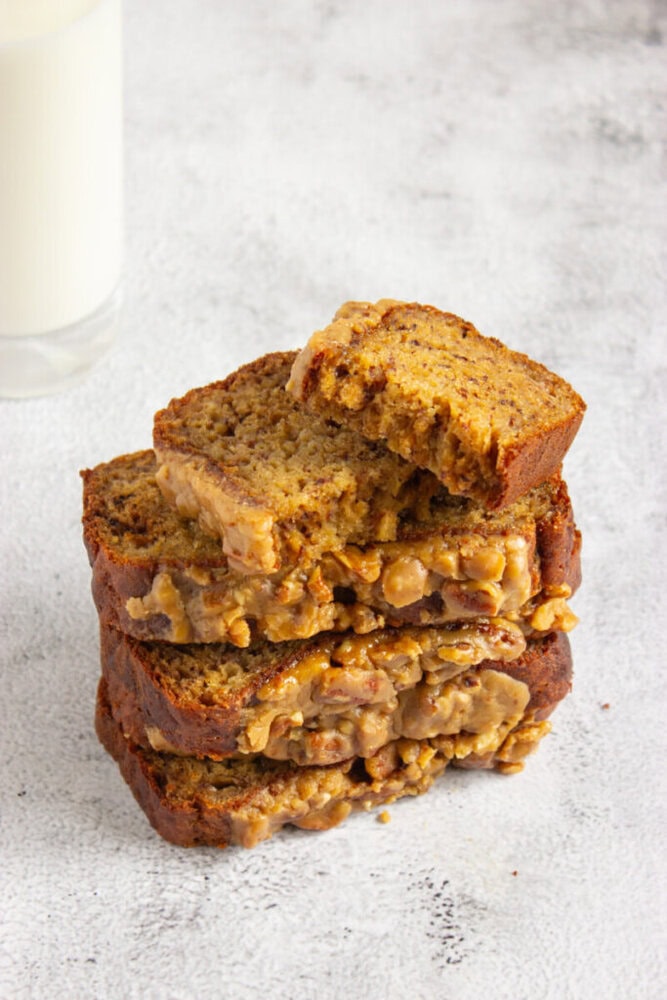 Four slices of banana bread stacked on top of each other, with oats and glaze, and a glass of milk in the background. The top slice has a bite taken out of it.