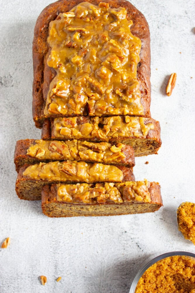 A loaf of bread topped with caramelized nuts, partially sliced, sits on a light surface with scattered pecans and a bowl of brown sugar nearby.