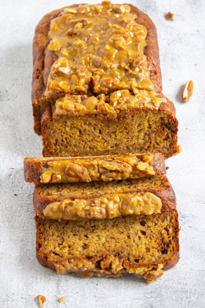 Sliced loaf of banana bread topped with a chopped walnut glaze, displayed on a light surface.