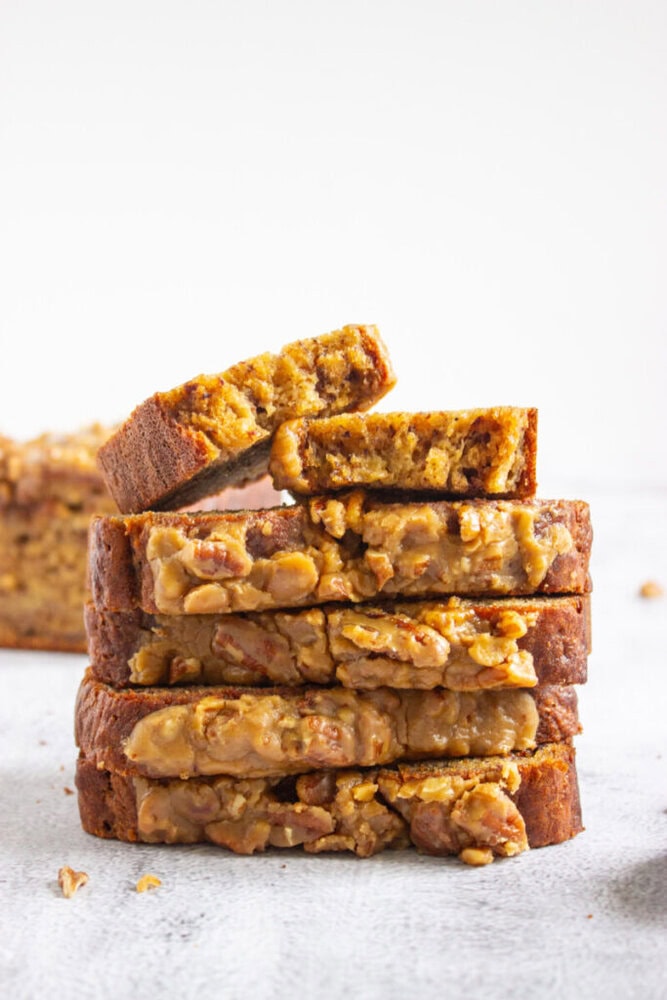 A stack of sliced banana bread with visible chunks of nuts, with the top slice slightly bitten, set on a light surface.