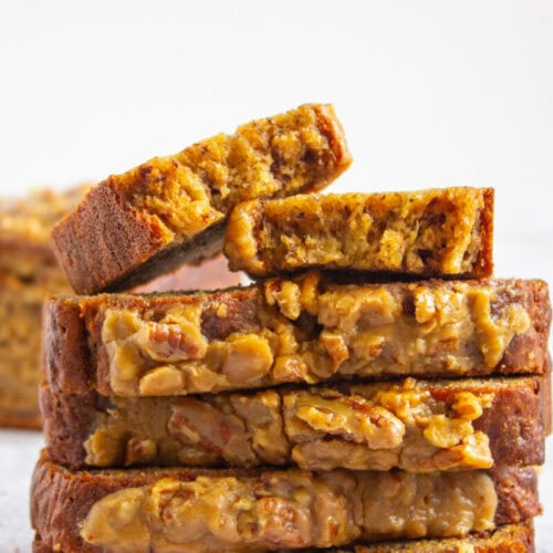 A stack of sliced banana bread with a nut topping, with one slice leaning on top, set against a plain white background.