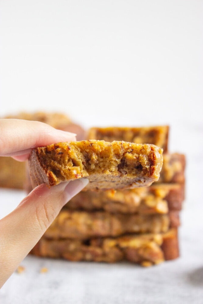 A hand holds a slice of banana bread with a bite taken out, with more slices stacked in the background.