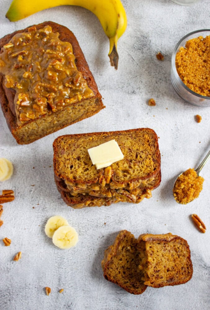 Sliced banana bread with pecan topping, a pat of butter on top, a whole banana, brown sugar, banana slices, and pecans on a light surface.