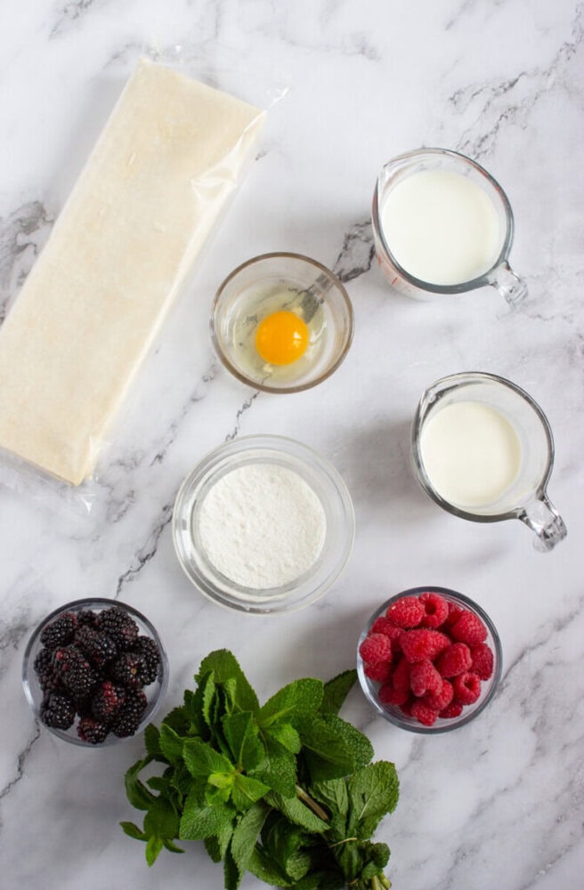 Ingredients on a marble surface: a block of pastry dough, an egg, two measuring cups of milk, a bowl of flour, bowls of blackberries and raspberries, and a bunch of fresh mint.
