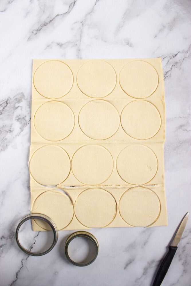 A sheet of dough with large circles cut out, a knife, and two round metal cutters on a marble surface.