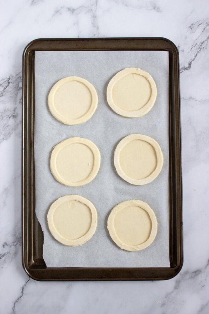 A baking tray lined with parchment paper holds six evenly spaced, unbaked round pastry shells on a marble surface.