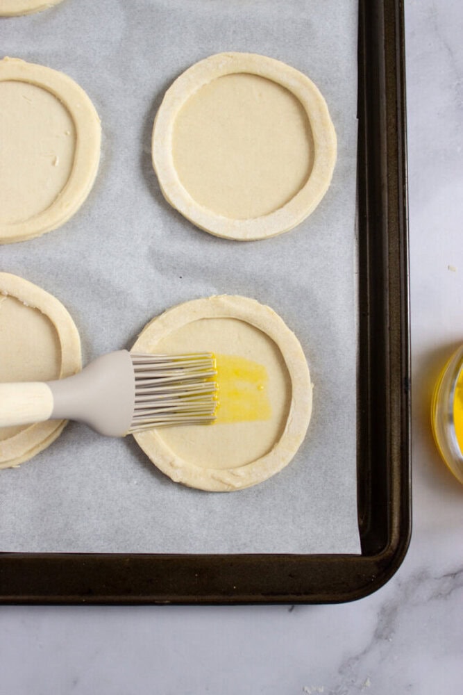 A pastry brush applies egg wash to a round piece of unbaked pastry dough on a parchment-lined baking sheet.