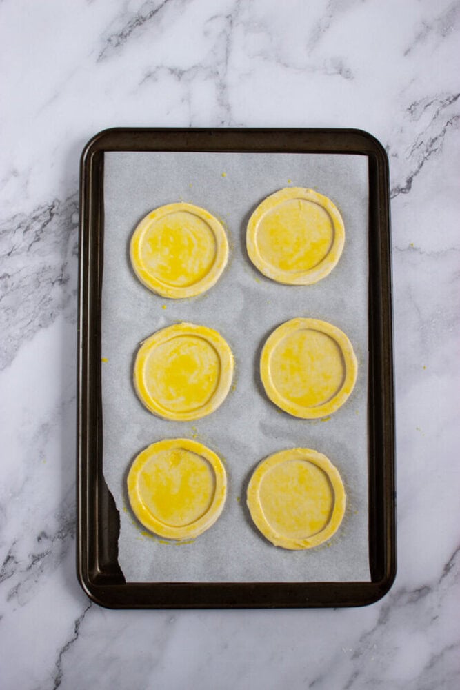 A baking tray lined with parchment paper holds six unbaked round pastry shells coated with an egg wash on a marble countertop.
