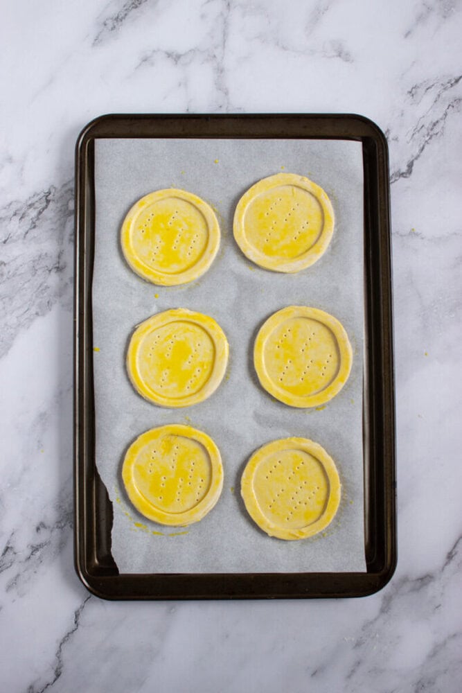 A baking sheet lined with parchment paper holds six circular, unbaked pastry shells brushed with egg wash, placed on a marble countertop.